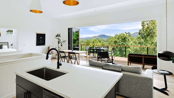 A modern kitchen with a large island, dining area with chairs, and a balcony with a view of greenery and mountains.