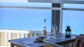 Une table en bois, avec une bouteille, des verres et une lanterne, sur un balcon donnant sur la mer.