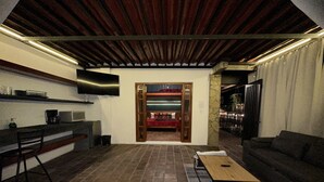 Una sala de estar moderna con piso de piedra, techo de madera, un televisor de pantalla plana montado en la pared, un sofá, una mesa de centro y un área de cocina con microondas y cafetera.