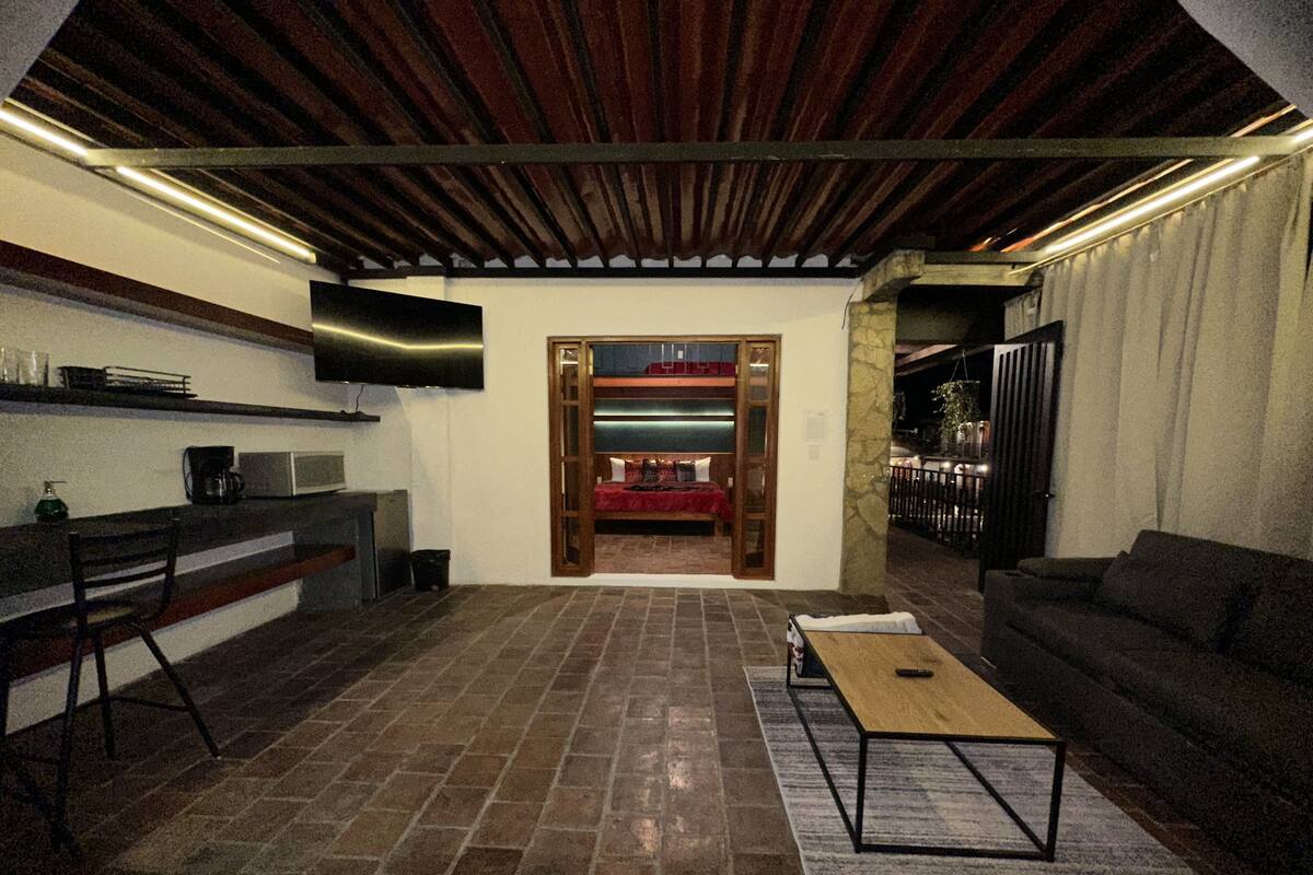 Un salon moderne avec un sol en pierre, un plafond en bois, un téléviseur à écran plat fixé au mur, un canapé, une table basse et un coin cuisine équipé d’un four à micro-ondes et d’une machine à café.