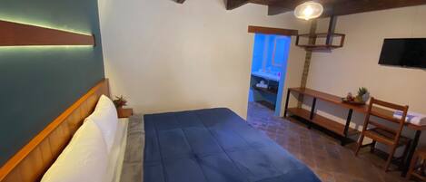 A bedroom with a bed, wooden headboard, a bench, a small table with fruit, and a TV on the wall.