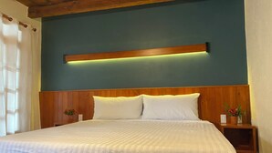 Una cama bien hecha con tablero de madera, una mesita de noche con un florero y una lámpara fijada a la pared.