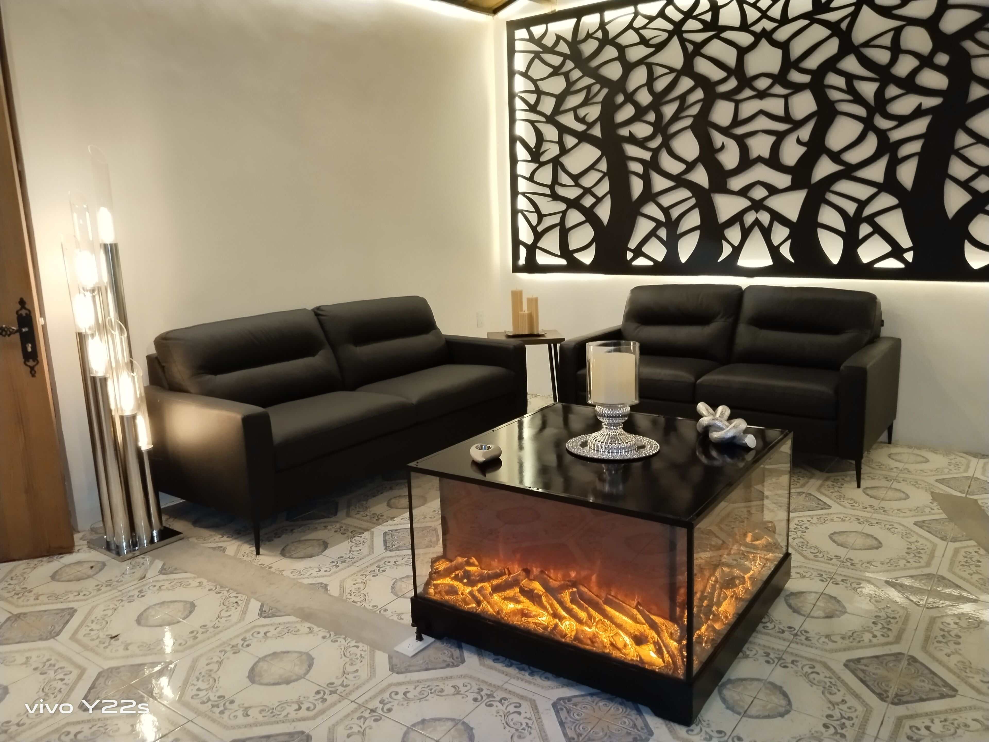 A modern living room with a glass-top coffee table, two black sofas, a decorative wall panel, and a fireplace.