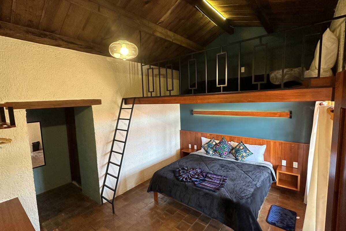 Une chambre à coucher compacte et rustique, avec un lit superposé, une échelle, une petite table de chevet et un lit recouvert d’une literie à motifs.