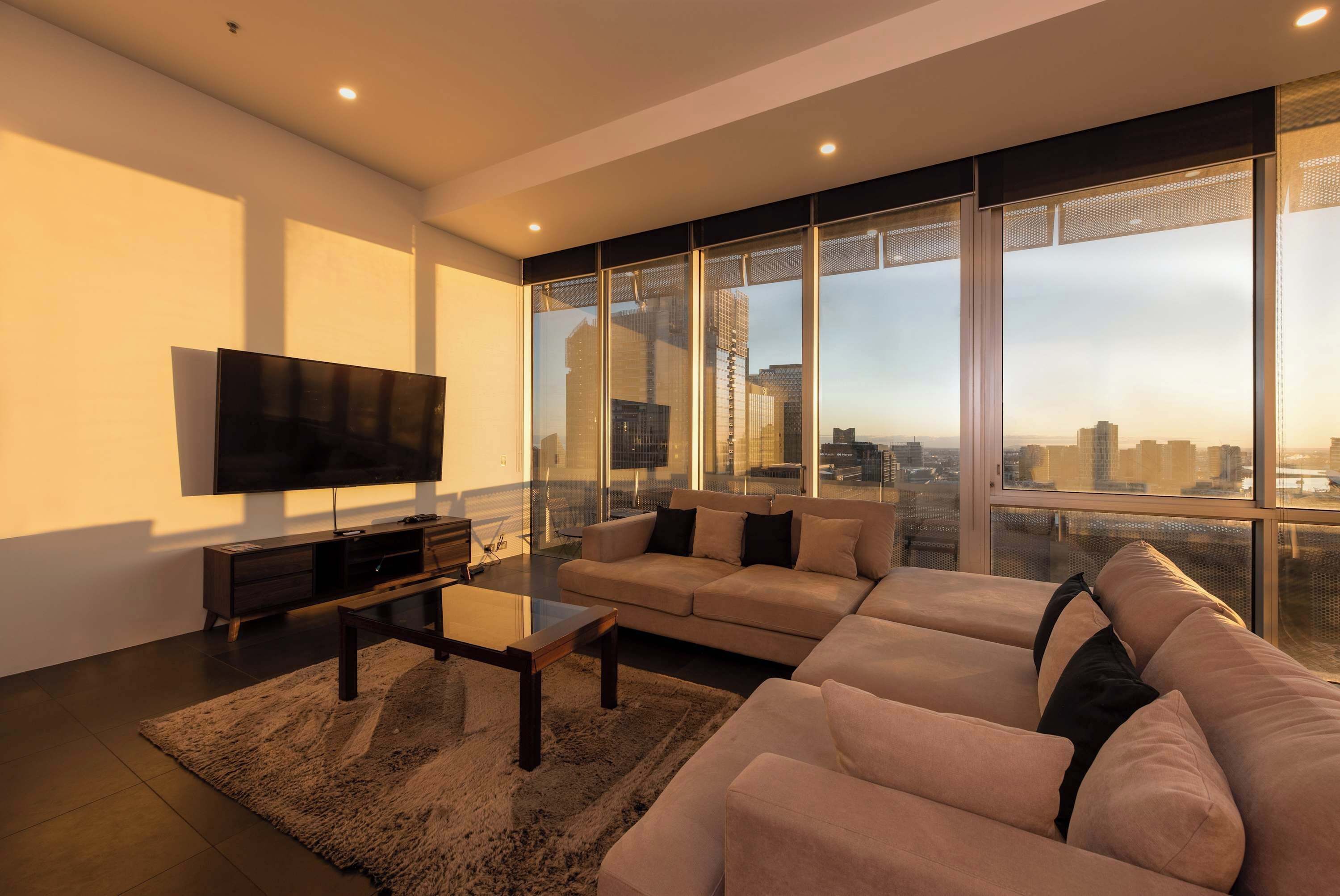 Een moderne woonkamer met een grote flatscreen televisie, een hoekbank en een stadsgezicht door de vloerhoogte ramen.