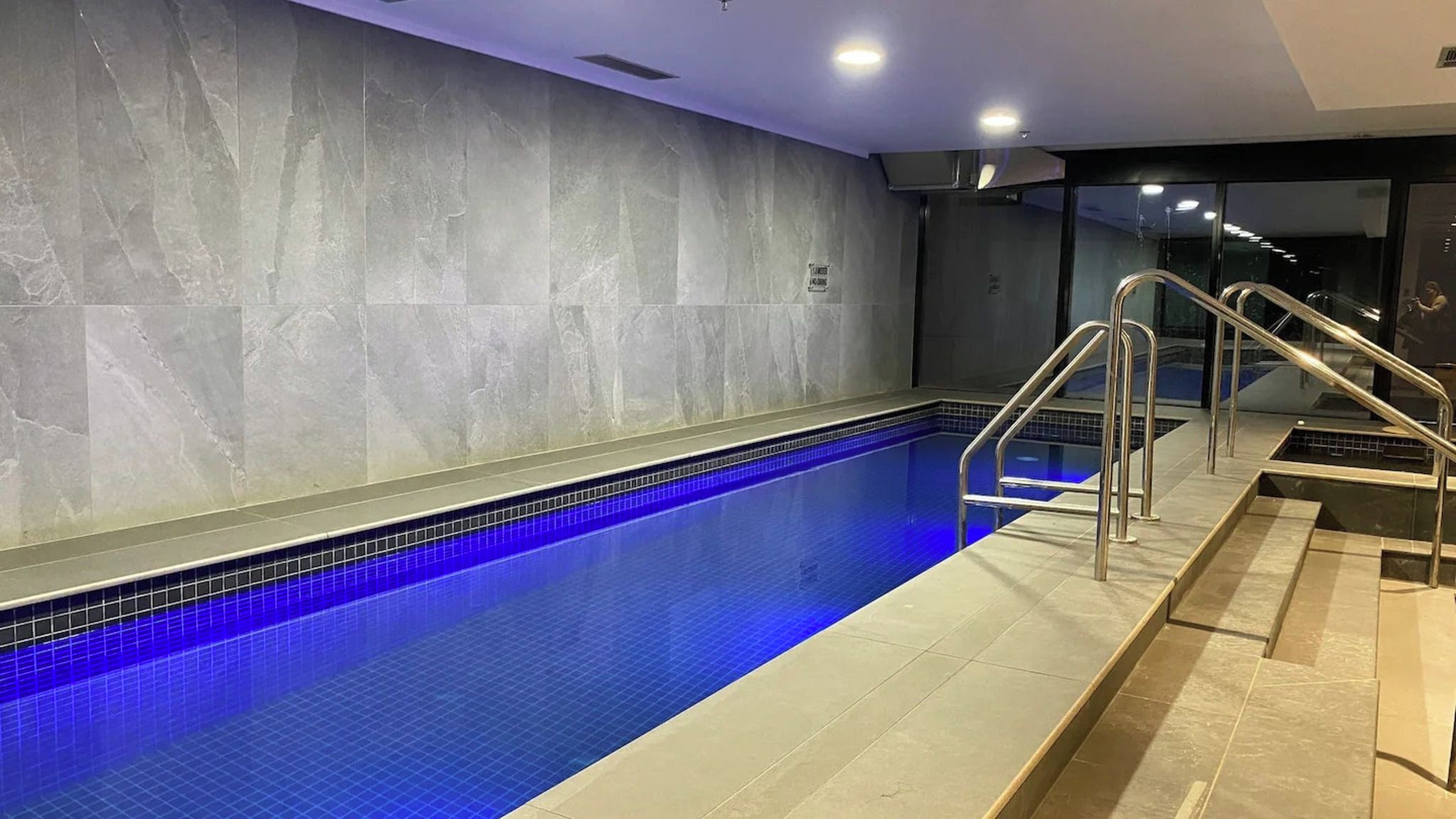 En swimmingpool med ett blått, kaklat botten, ett metallräcke och en kaklad vägg med en strukturerad yta.