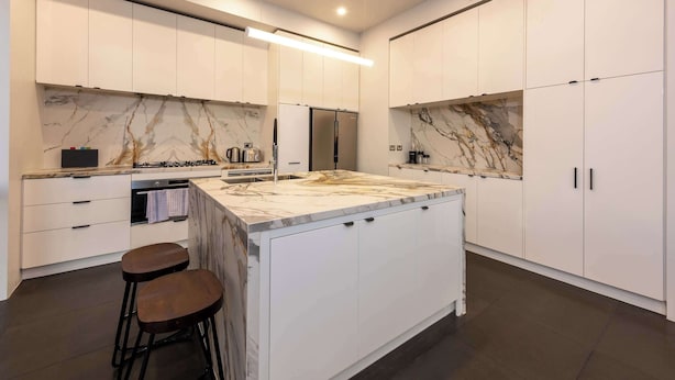 A modern kitchen with white cabinetry, a marble countertop, and a built-in island with stools. - Spacious CBD Penthouse w Parking Pool and Gym (West Melbourne)