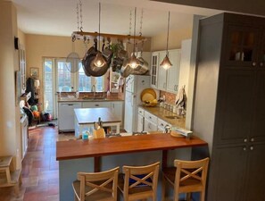A kitchen with a central island, wooden chairs, hanging pendant lights, and a view of the adjacent living area.