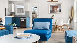 En moderne stue med en blå sofa, et rundt sofabord med bøger, et spiseområde med et hvidt bord og stole, og et køkken i baggrunden.