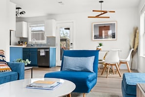 Ein modernes Wohnzimmer mit einem blauen Sofa, einem runden Couchtisch mit Büchern, einem Essbereich mit weißem Tisch und Stühlen und einer Küche im Hintergrund.