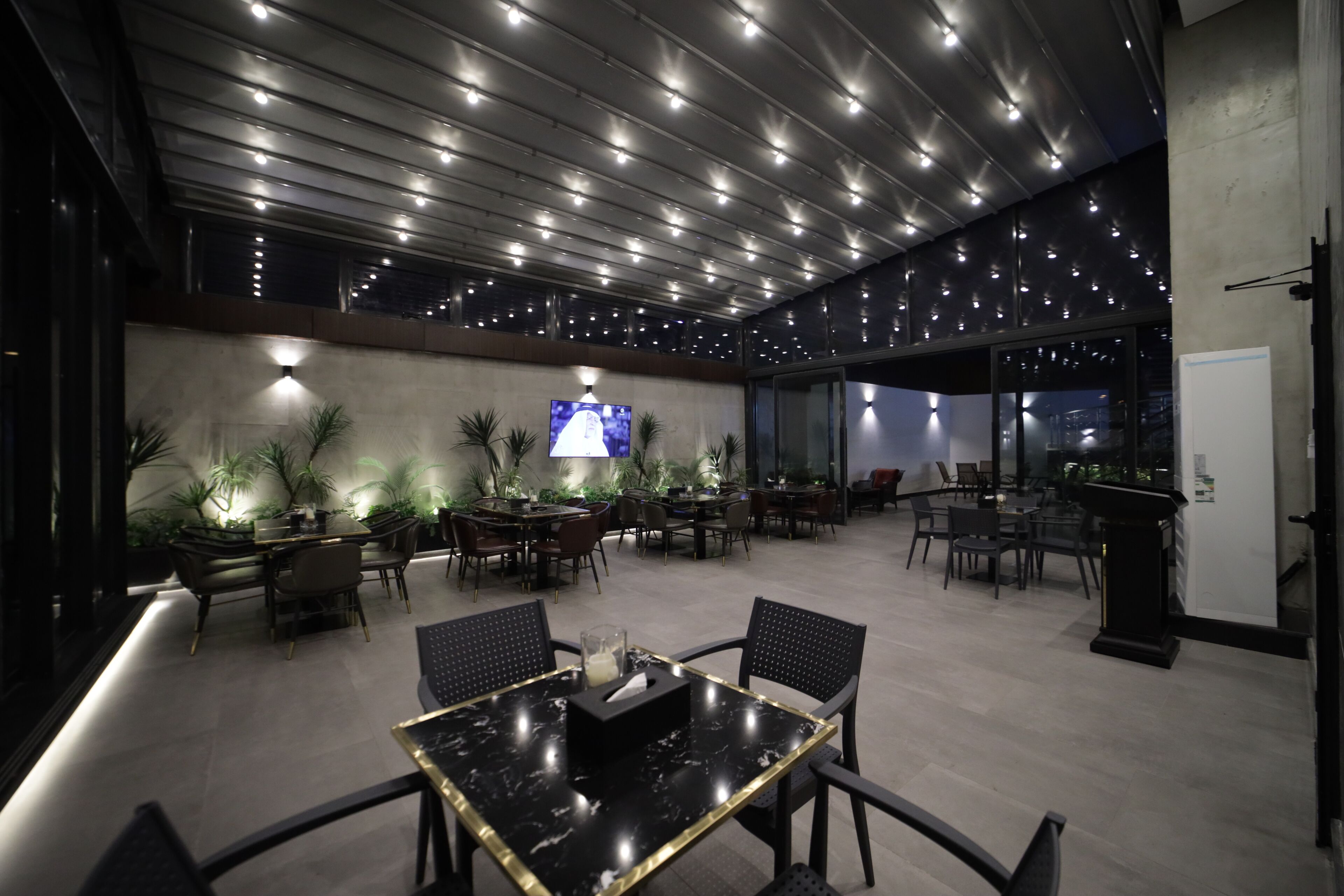 Un espace repas moderne avec une table en marbre noir, plusieurs tables à manger, des chaises, des plantes en pot et un écran de télévision.