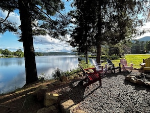 En sittplats vid sjön med Adirondack-stolar och ett litet bord, omgiven av träd och med utsikt över sjön.