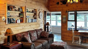 L’intérieur chaleureux d’un chalet avec un plafond en bois, un ventilateur de plafond, un canapé en cuir marron, un tapis à motifs et une fenêtre donnant sur le paysage extérieur.