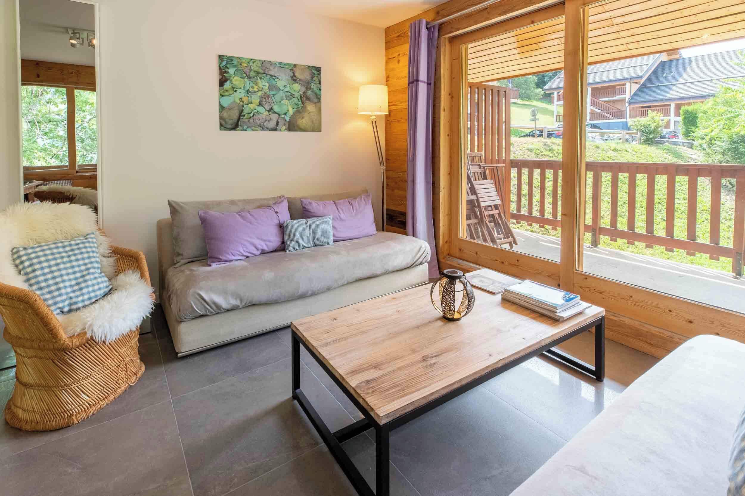 Uma sala de estar acolhedora com um sofá, uma poltrona de vime, uma mesa de centro de madeira e vista para um deck de madeira e vegetação lá fora.