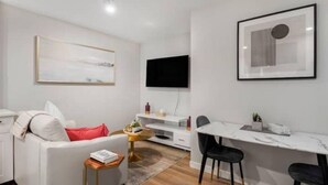 Ett modernt vardagsrum med en vit soffa, en röd kudde, ett litet sidobord, ett vitt skrivbord med en stol och en platt-TV som är monterad på väggen.