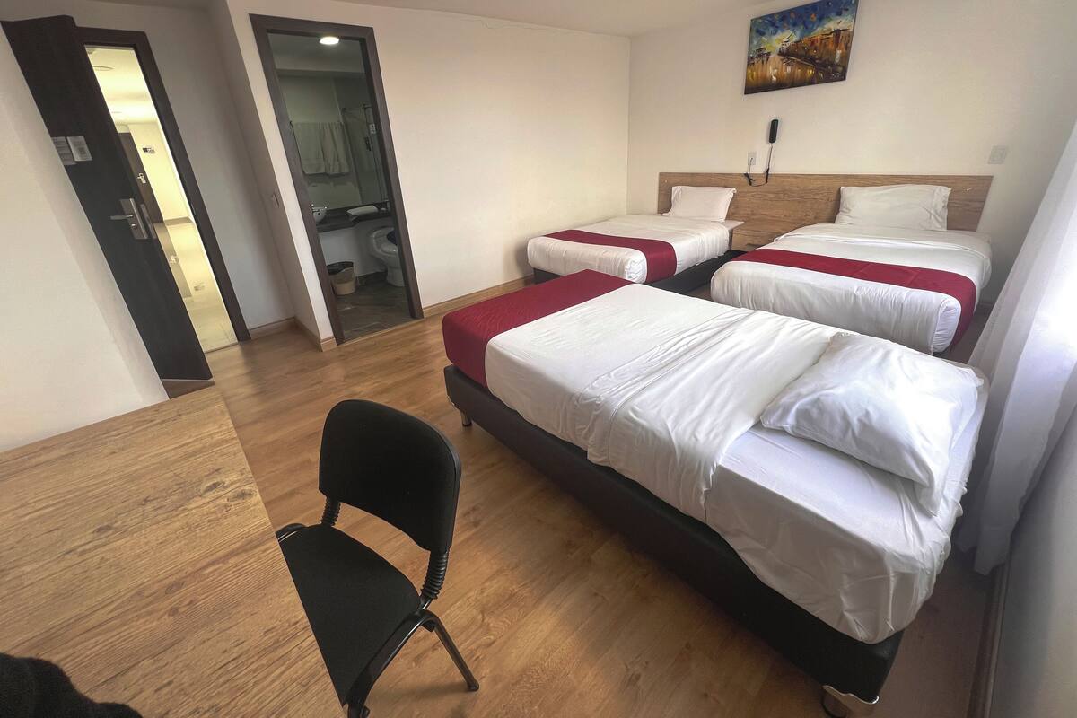A hotel room with three beds, a wooden floor, a black chair, and a painting on the wall.