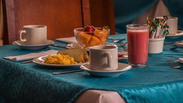 Un desayuno con una mesa cubierta por un mantel azul, que incluye un plato de huevos revueltos, tostadas, un frutero con melón y fresas, un vaso de batido rosa y una taza blanca.
