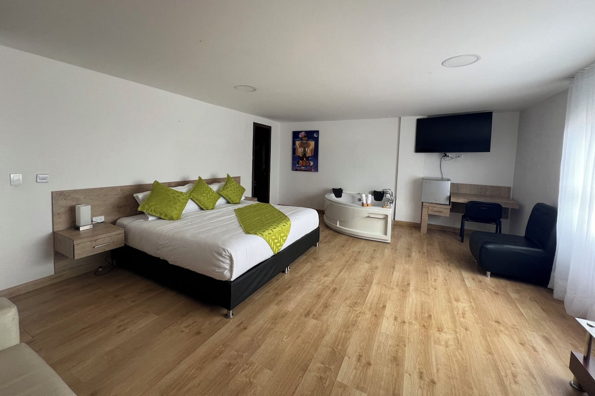 A modern hotel room with a large bed, a desk, a chair, a TV, and a bathtub.