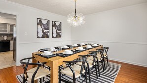 A dining area with a long wooden table set for six, surrounded by black chairs with woven seats. Above the table, there are two abstract paintings on the wall. - Hot tub, Fire-pit, Pool Table, Heated Pool (Jamestown)