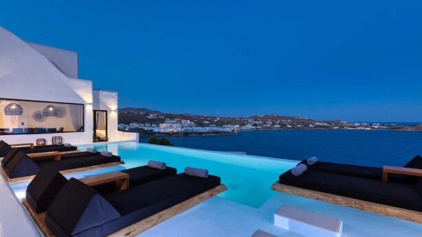 Ein moderner Infinity-Pool mit Liegestühlen und Blick auf eine Küstenstadt im Abendlicht.