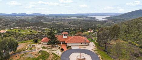 A villa with a red-tiled roof, surrounded by a landscaped garden and a view of a valley and mountains.