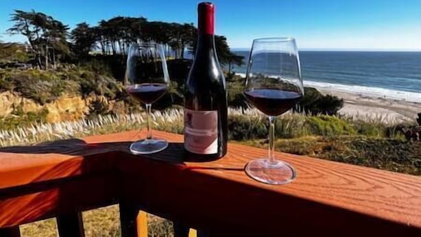 Une table en bois, une bouteille de vin et deux verres à vin sont posés sur un balcon donnant sur l’océan.