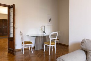 Un comedor con una mesa de vidrio, dos sillas blancas con cojines amarillos, una planta en maceta y un cuadro de un búho en la pared.