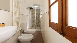 Ein schmales Badezimmer mit einem weißen Waschbecken, zwei Toiletten, einem Bidet und einer Duschkabine aus Glas.