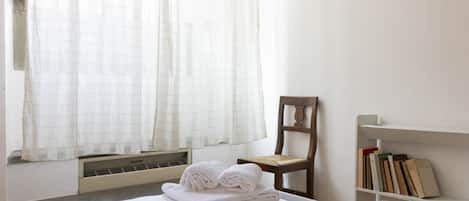 Habitación de hotel con cama, una silla, una estantería y una ventana con cortinas.