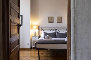 Un dormitorio con puerta de madera, mesita de noche con lámpara, cama con almohadas y dos cuadros en la pared.