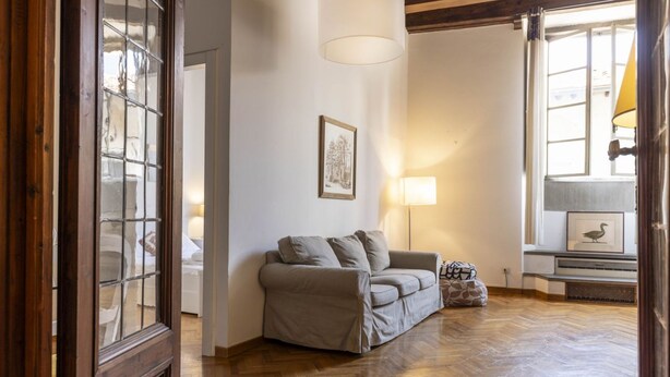 Ein gemütliches Wohnzimmer mit Holzdielen im Fischgrätmuster, einem beigen Sofa, einer Lampe und einem gerahmten Bild an der Wand.