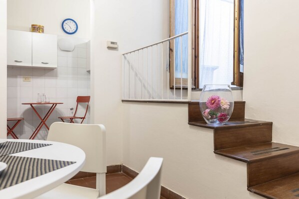 A modern kitchen with white cabinets, a dining table with chairs, a clock, and a staircase with a vase of flowers.