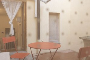 A small patio with a round table and two chairs, a window with a blind, and a door with a curtain.