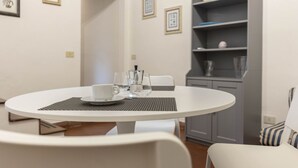 A round dining table with a white tablecloth, a white chair, and a small white table lamp. There is a bookshelf with various items on the shelves. - Casa Florentia - Florence (Firenze)