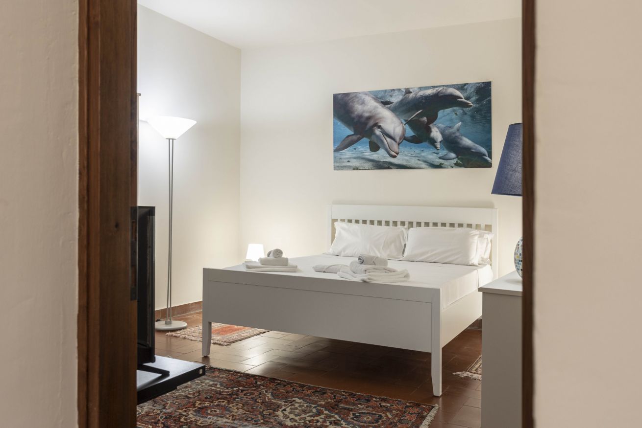 A bedroom with a bed, a nightstand, a lamp, a painting of dolphins, and a patterned rug.