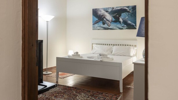 A bedroom with a bed, a nightstand, a lamp, a painting of dolphins, and a patterned rug. - Casa Florentia - Florence (Firenze)