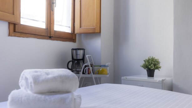 Quarto bem arrumado com cama, cafeteira, mesinha com planta e janela com persianas de madeira.