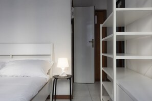Et hotelværelse med en seng, et natbord med lampe samt et hvidt skab med hylder.