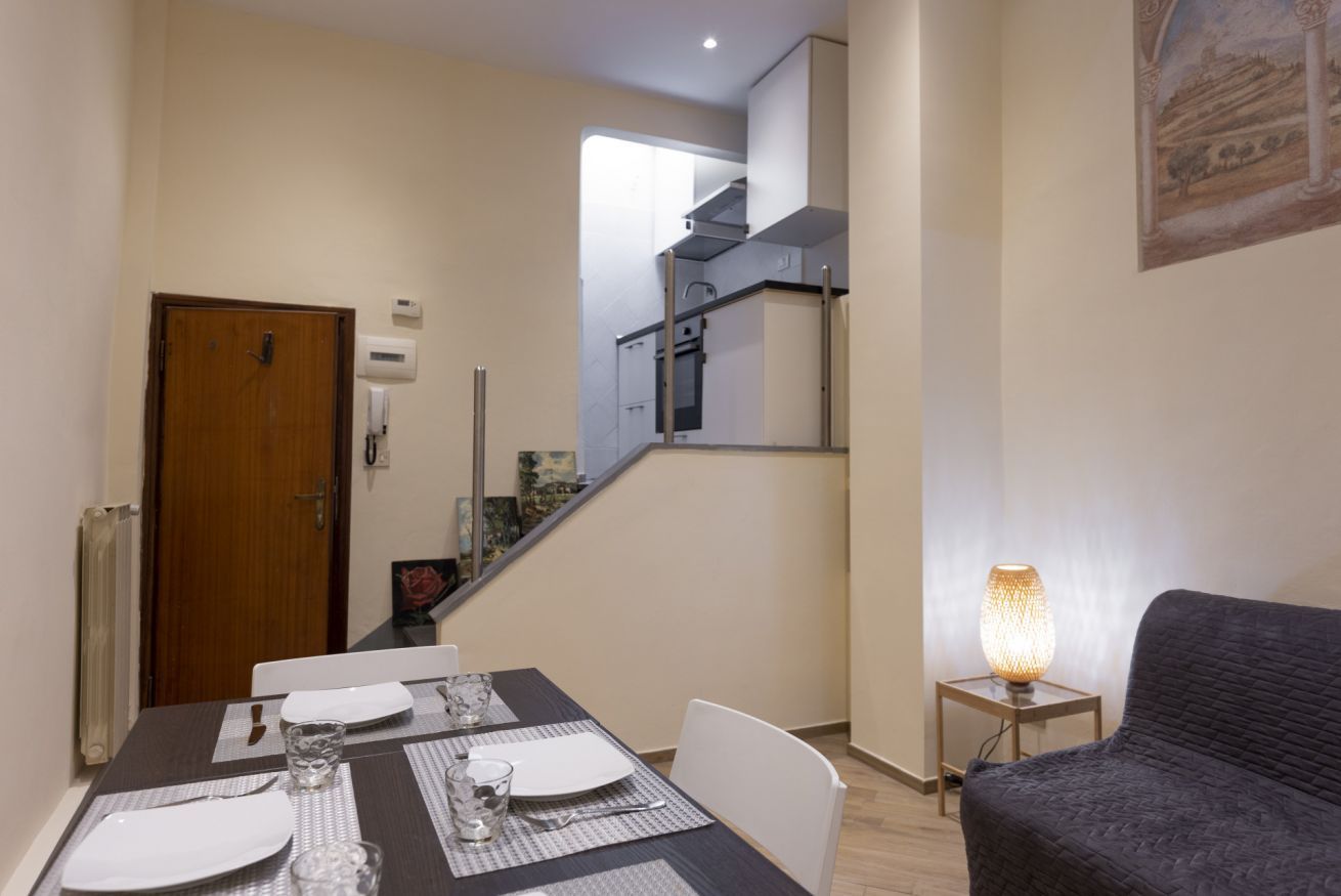 Un comedor con una mesa dispuesta para cuatro, una escalera que lleva a una cocina, y una lámpara sobre una mesita auxiliar.