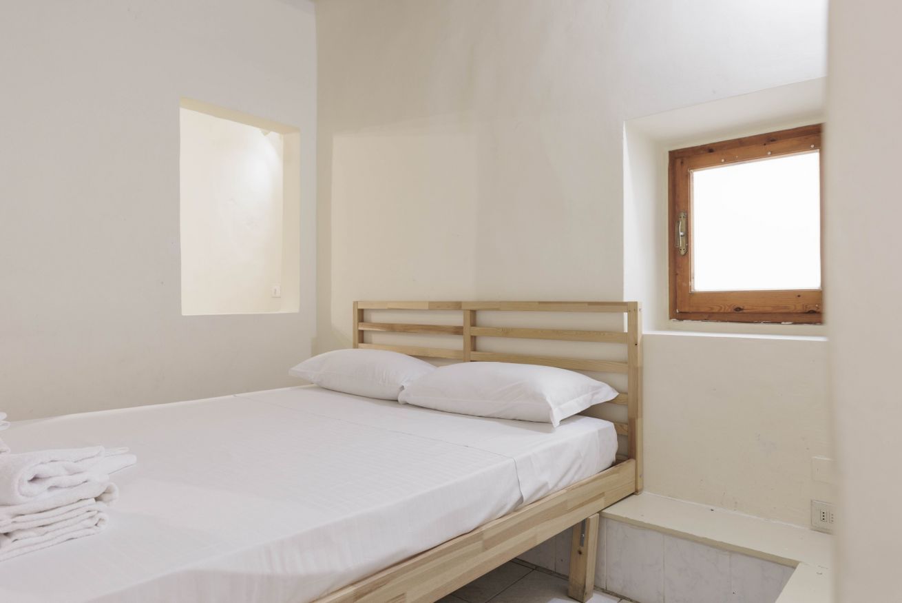 A small, single-bed room with a wooden headboard, a window, and a small bench or table.