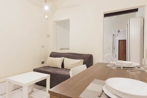 Une pièce moderne et bien éclairée, avec un canapé marron, une table blanche et un coin repas comprenant une table en bois et de l’assiette blanche.