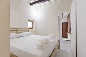 Une chambre avec une tête de lit en bois, un lit recouvert de draps blancs et une petite fenêtre. Des serviettes sont pliées sur le lit et un banc en bois se trouve dans un coin.
