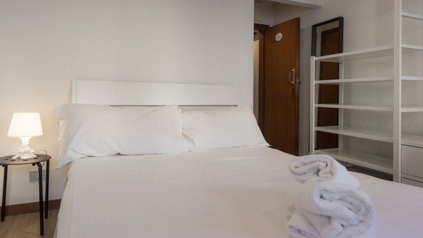 Habitación de hotel con cama, mesita de noche con lámpara, puerta de madera y estante blanco.