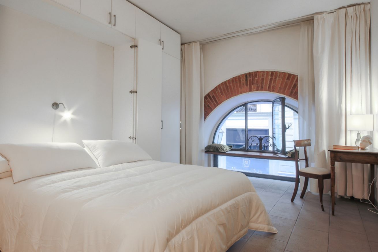 Une chambre avec un grand lit, un bureau et une vue par une fenêtre surmontée d’une arche en briques.