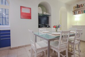 Un coin repas avec une table en verre, des chaises blanches, un téléviseur à écran plat fixé au mur et une bibliothèque remplie de livres.
