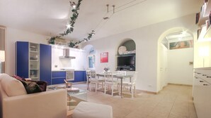 A modern living room with a sofa and ottoman, a dining area with a table and chairs, a kitchen with blue cabinets and a TV, and a hallway with an archway. - Duomo Deluxe Apartment (Firenze)