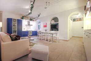 Un salon moderne avec un canapé et un pouf, un coin repas avec une table et des chaises, une cuisine avec des meubles bleus et un téléviseur, et un couloir avec une arche.