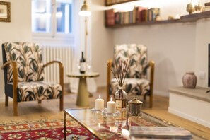 A living room with two patterned armchairs, a glass coffee table with candles, a vase, and a radiator in the background.
