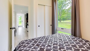 A bedroom with a large window, a bed with a patterned bedspread, and a view of a green outdoor area. - Lakefront Catskills Container Home: Private Lake (Swan Lake)