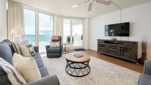 A spacious living room with a large window overlooking the ocean, a ceiling fan, a flat-screen TV, and a wooden coffee table. - OE at White Surf Unit 306 (Daytona Beach Shores)